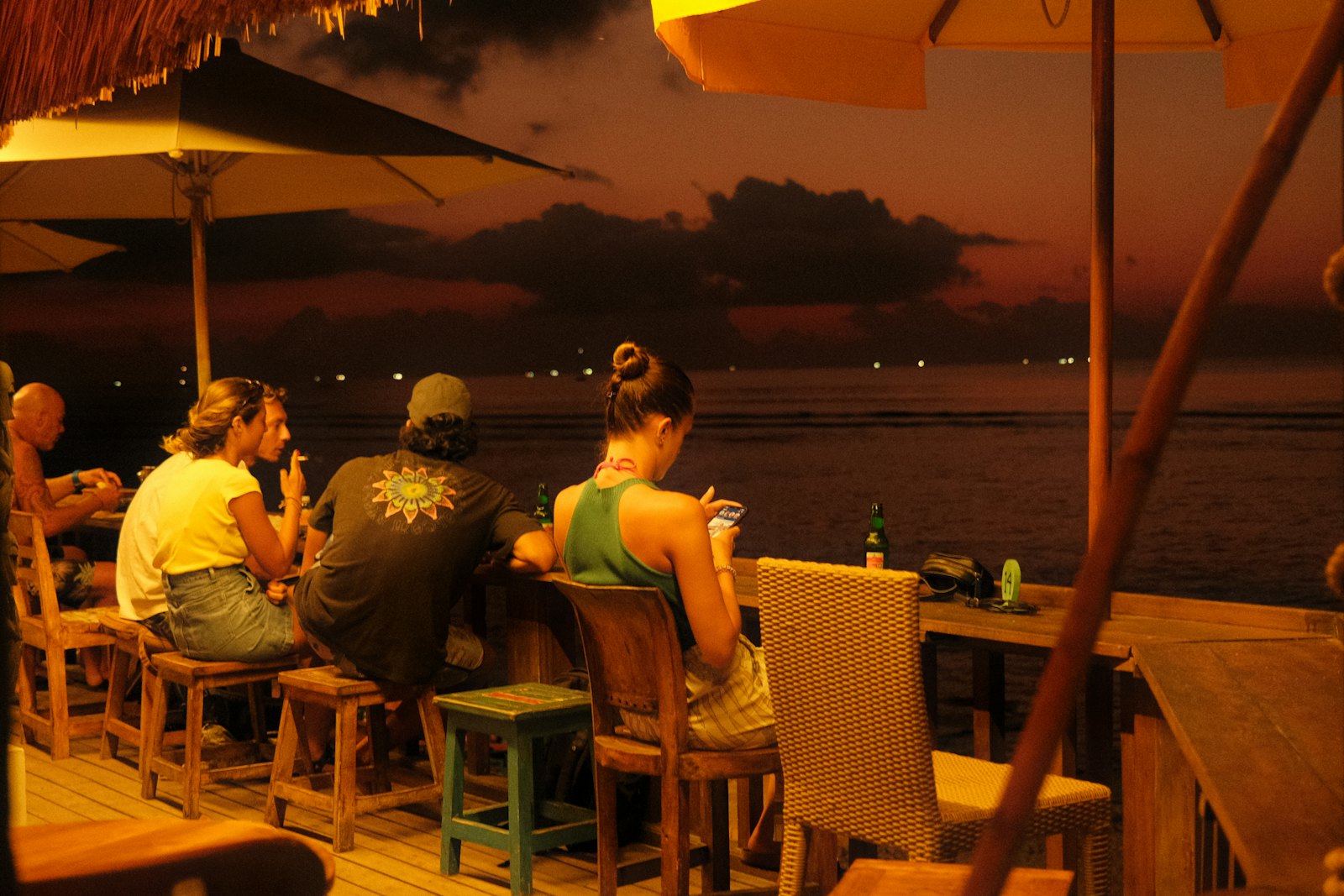 Surfers at a thatched beach bar at dusk, looking out at the water